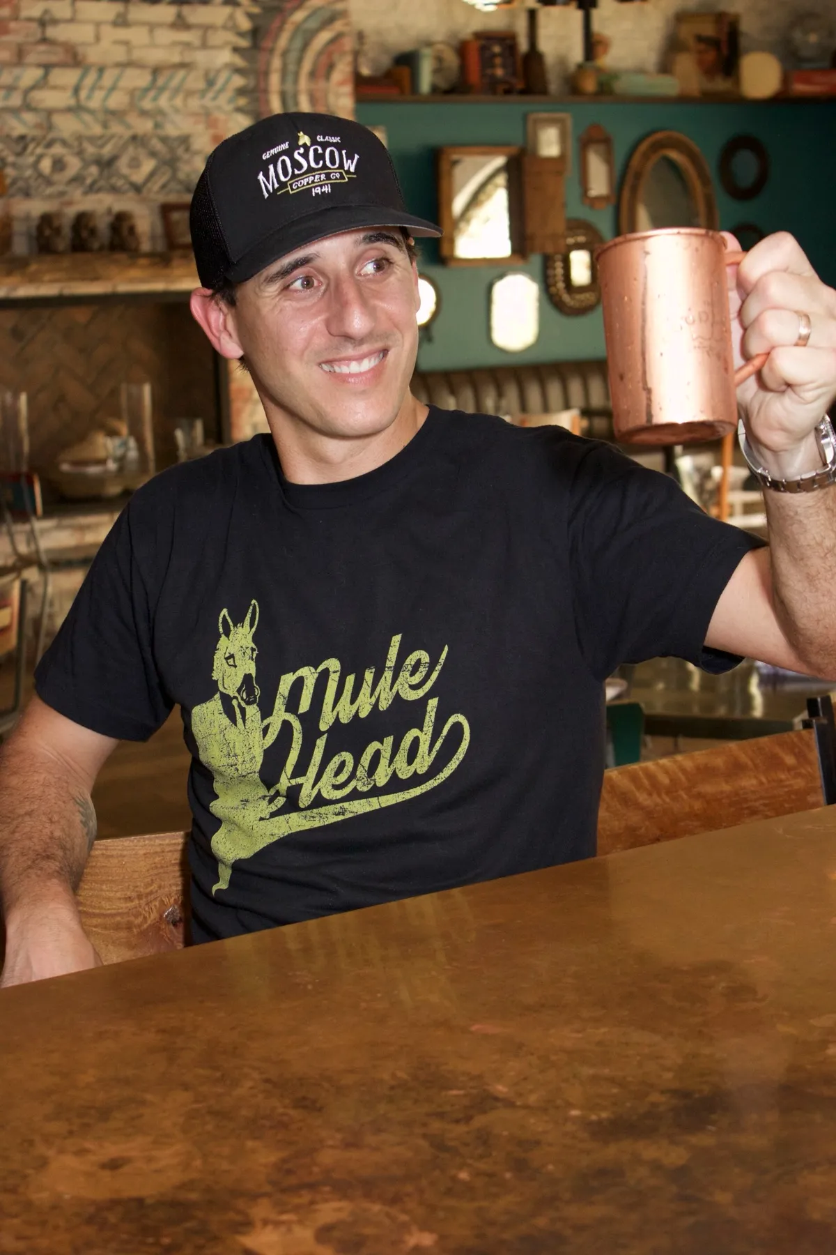 JJ Resnick behind a copper-topped bar in a Moscow Copper Co. cap and Mule Head shirt, raising a copper mug.