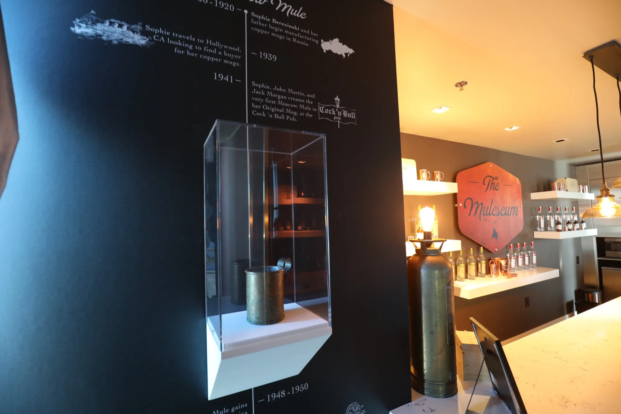 The original 1941 copper mug, heavily oxidised, in an acrylic vitrine against a wall printed with a dated Moscow Mule timeline.