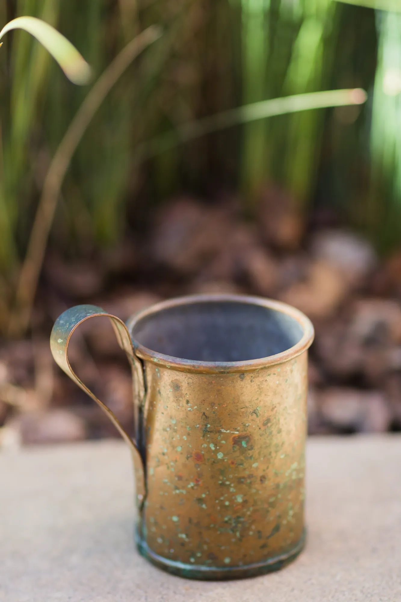 The original copper mug, oxidised to green, standing on gravel with its rim, handle and base visible.