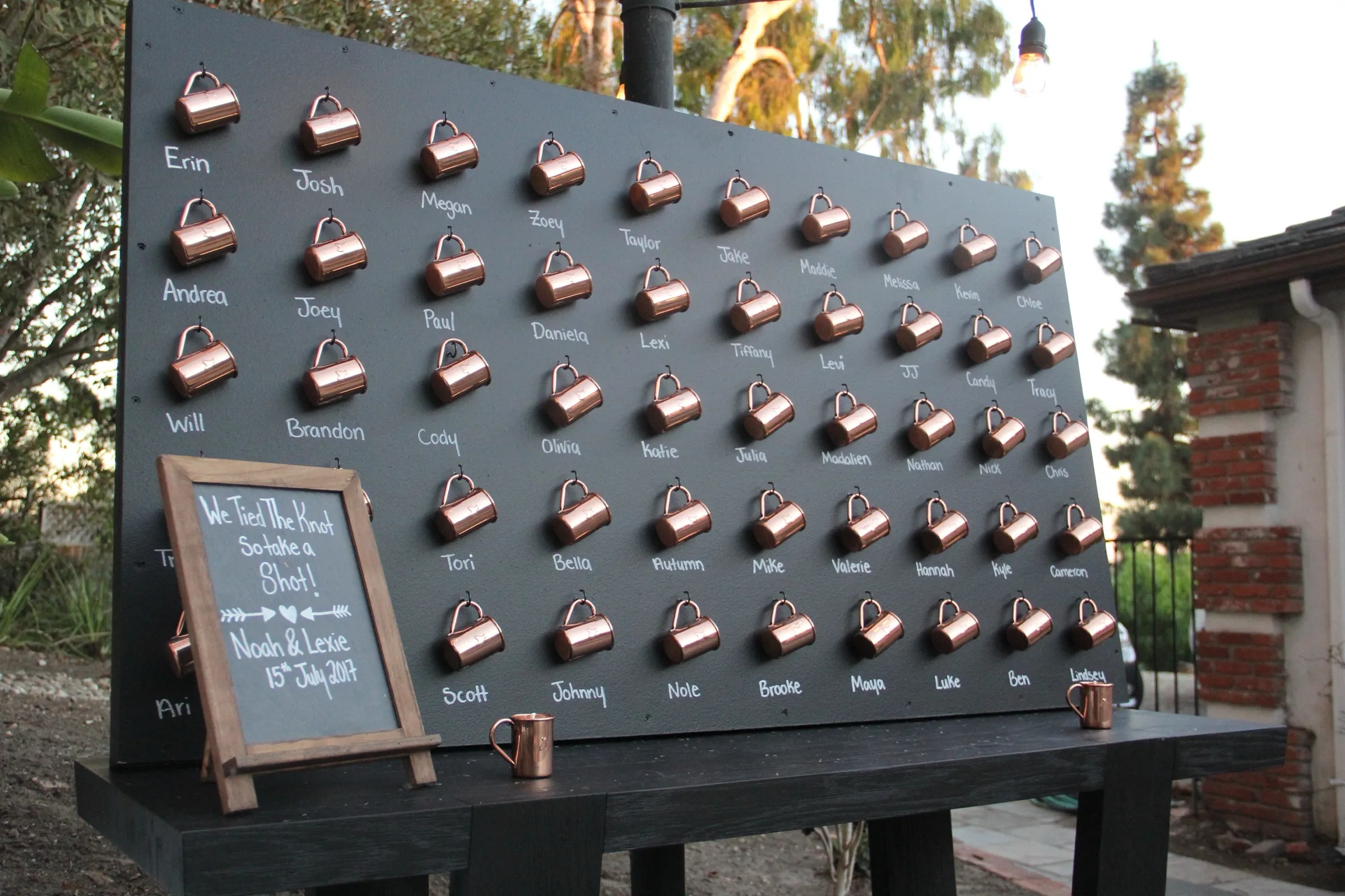 A freestanding slate wall hung with dozens of numbered copper mugs, each hand-lettered with a guest name.