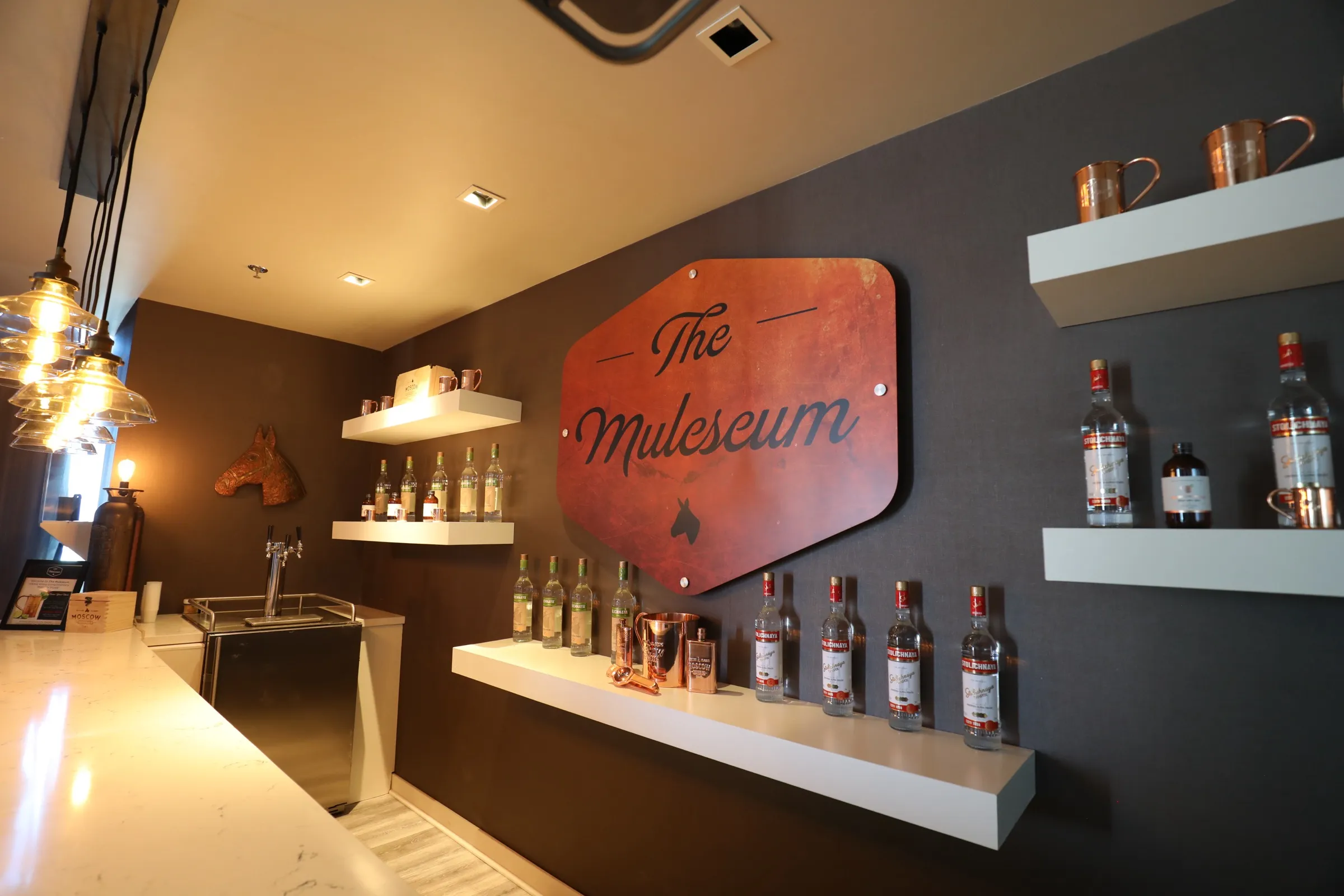 A wall at the Muleseum: copper mugs on a high shelf, bottles below, and a rust-coloured Muleseum sign with a brass mule-head sconce.