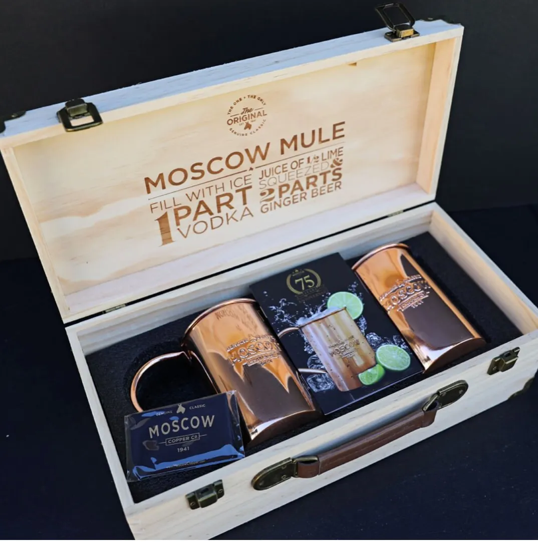 An open wooden two-pack presentation box: an engraved recipe in the lid, die-cut foam, two copper mugs, a card set, a leather handle and brass latches.