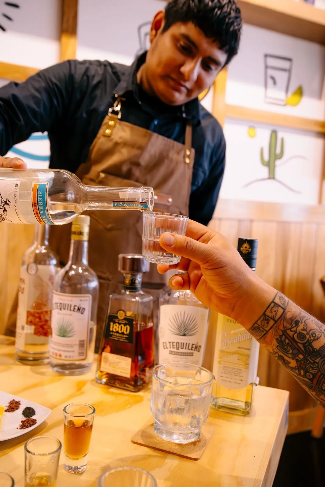 A bartender at Agavia pouring tequila into a tasting glass held out by a customer, with the flight of bottles on the counter.