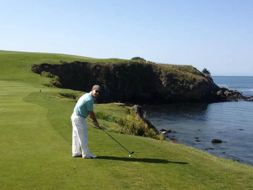 A golfer addressing the ball on a cliff-top tee above the Pacific, the fairway and the rocky shoreline behind.