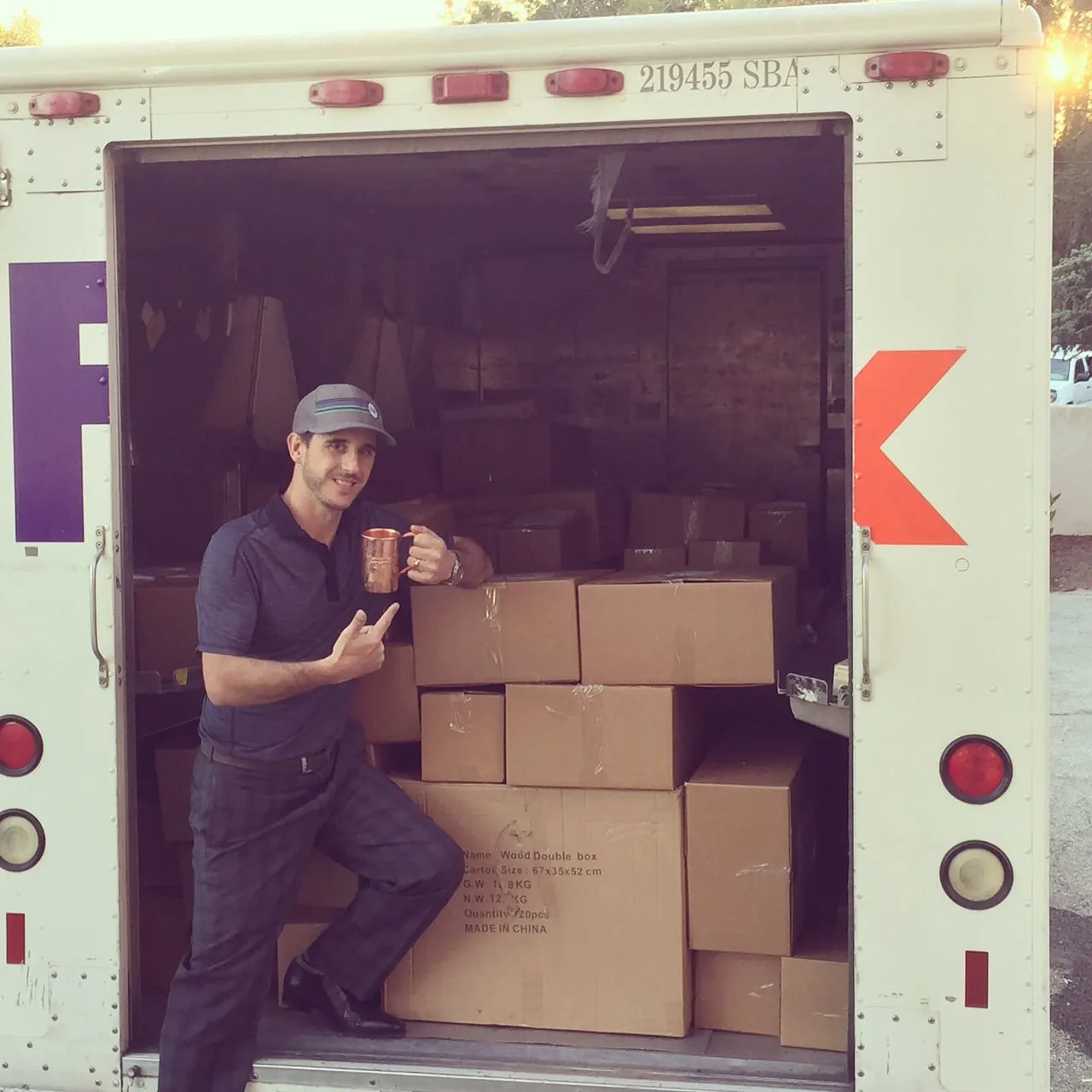 JJ Resnick standing in the back of a delivery truck beside stacked cartons, holding a copper mug.